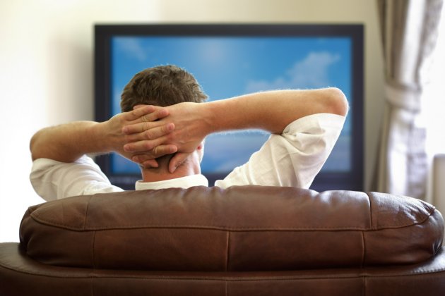 Man sitting on a sofa watching tv with hands folded behind his head